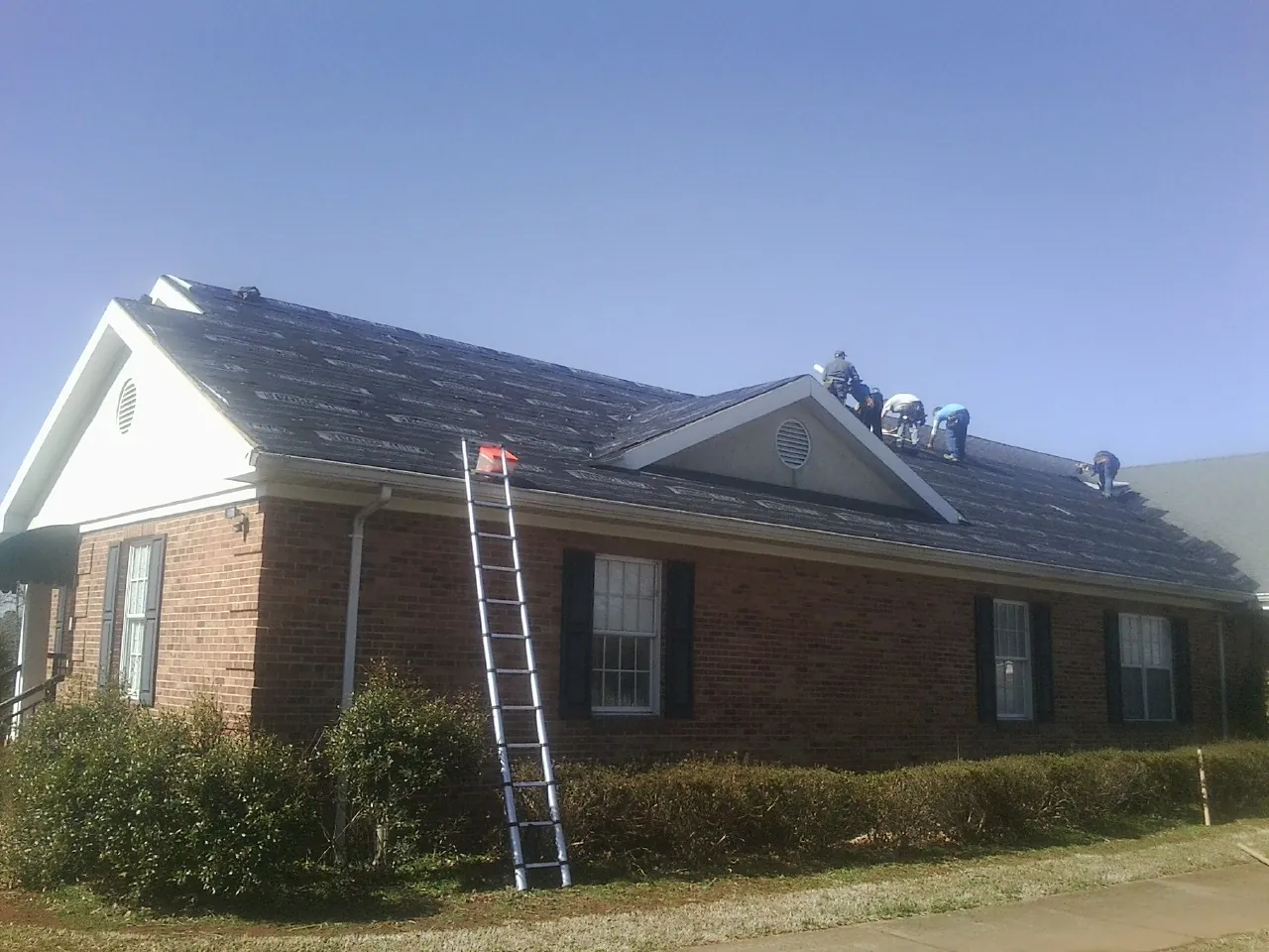 Crew tearing shingles off a brick ranch roof with a ladder set — Emergency Roof Repair work by our crew in Deforest