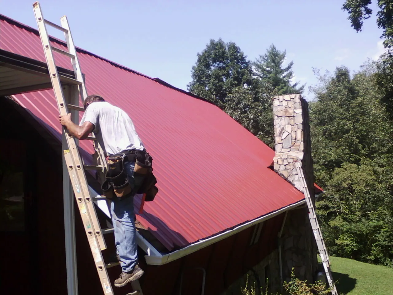 Roofer on a ladder beside a red metal roof and stone chimney — Soffit & Fascia Repair by Roofing Brothers in Deforest